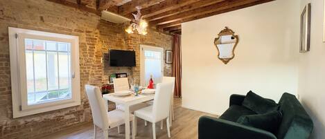 A dining area with a brick wall, wooden beams, a chandelier, a flat-screen TV, and a green sofa.