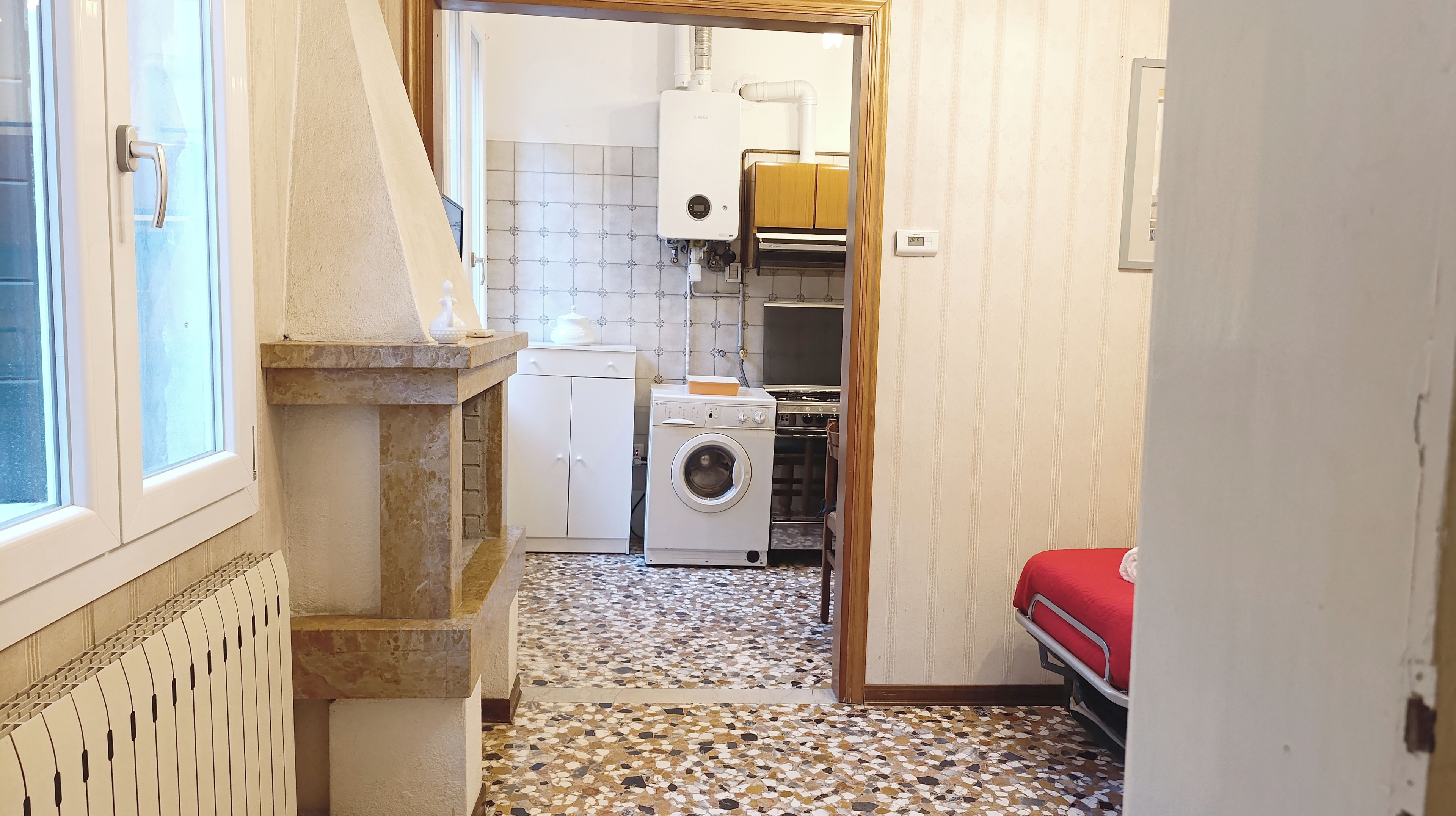 A narrow hallway with a radiator, a washing machine, and a bed with a red cover.