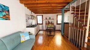 A cozy living area with a sofa, a dining table with chairs, a kitchen with a window, and a staircase.