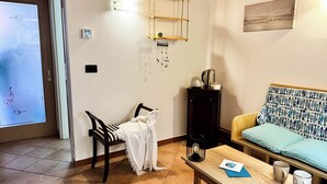 A cozy living room with a sofa, a coffee table, and a chair. There is a wall-mounted shelf with a kettle and a small table. A welcome sign hangs on the wall. - Di Sole e D'azzurro by Ca-sa Check (Chioggia)