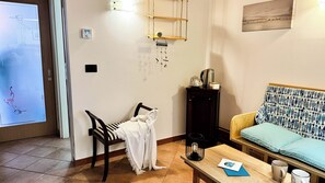 A cozy living room with a sofa, a coffee table, and a chair. There is a wall-mounted shelf with a kettle and a small table. A welcome sign hangs on the wall. - Di Sole e D'azzurro by Ca-sa Check (Chioggia)