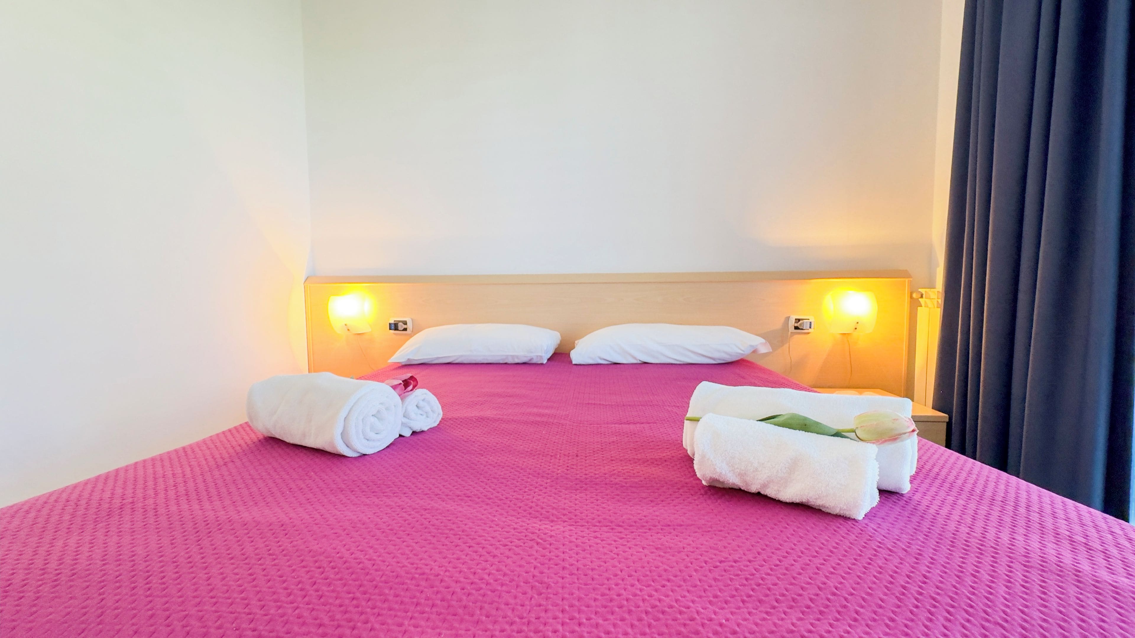 Ein Hotelzimmer mit einem rosa strukturierten Bettüberwurf, zwei weißen Kissen und zwei gerollten Handtüchern auf dem Bett.