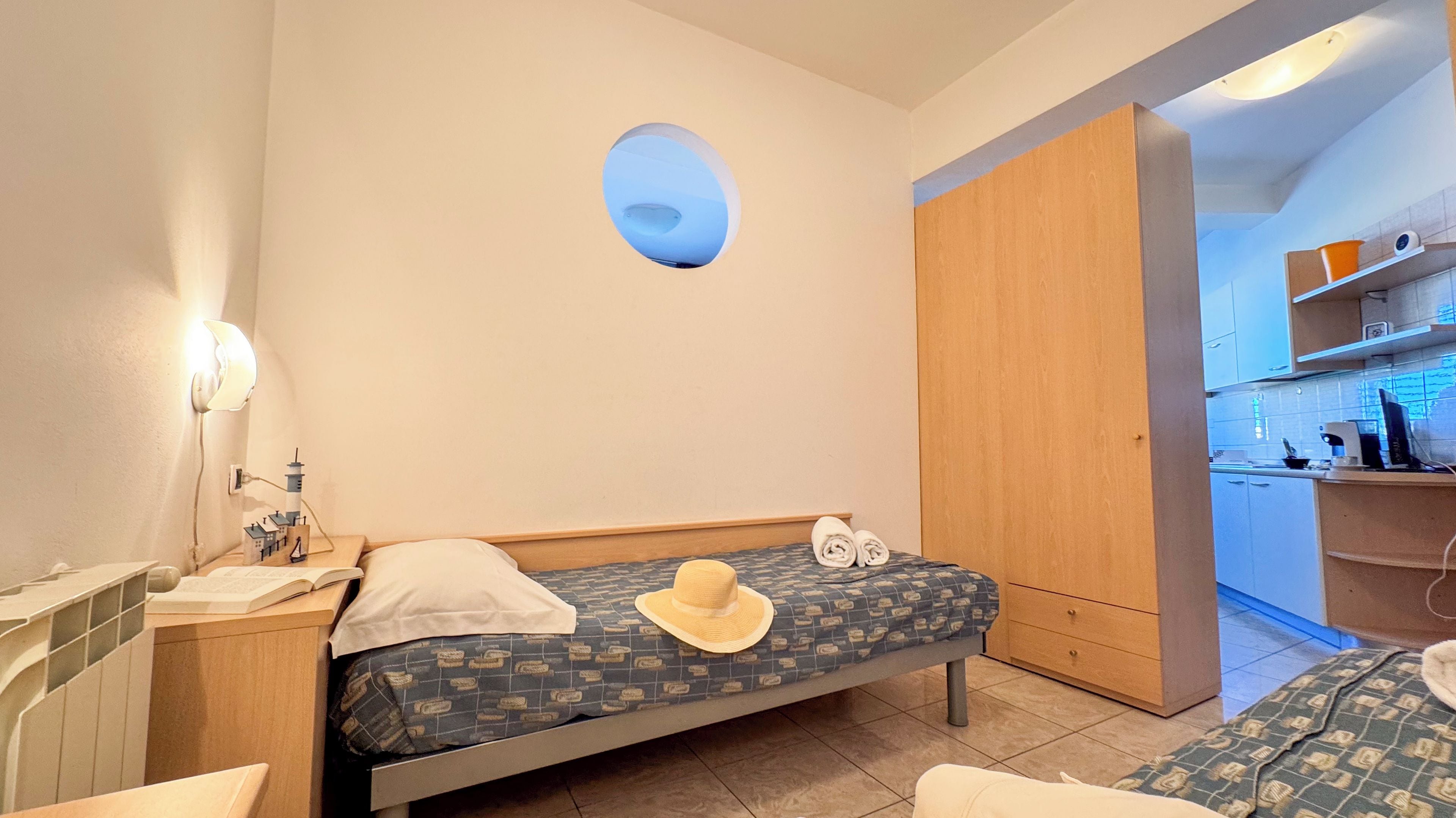 A compact bedroom with a bed, a bedside table, a lamp, a small window, and a kitchenette in the background.