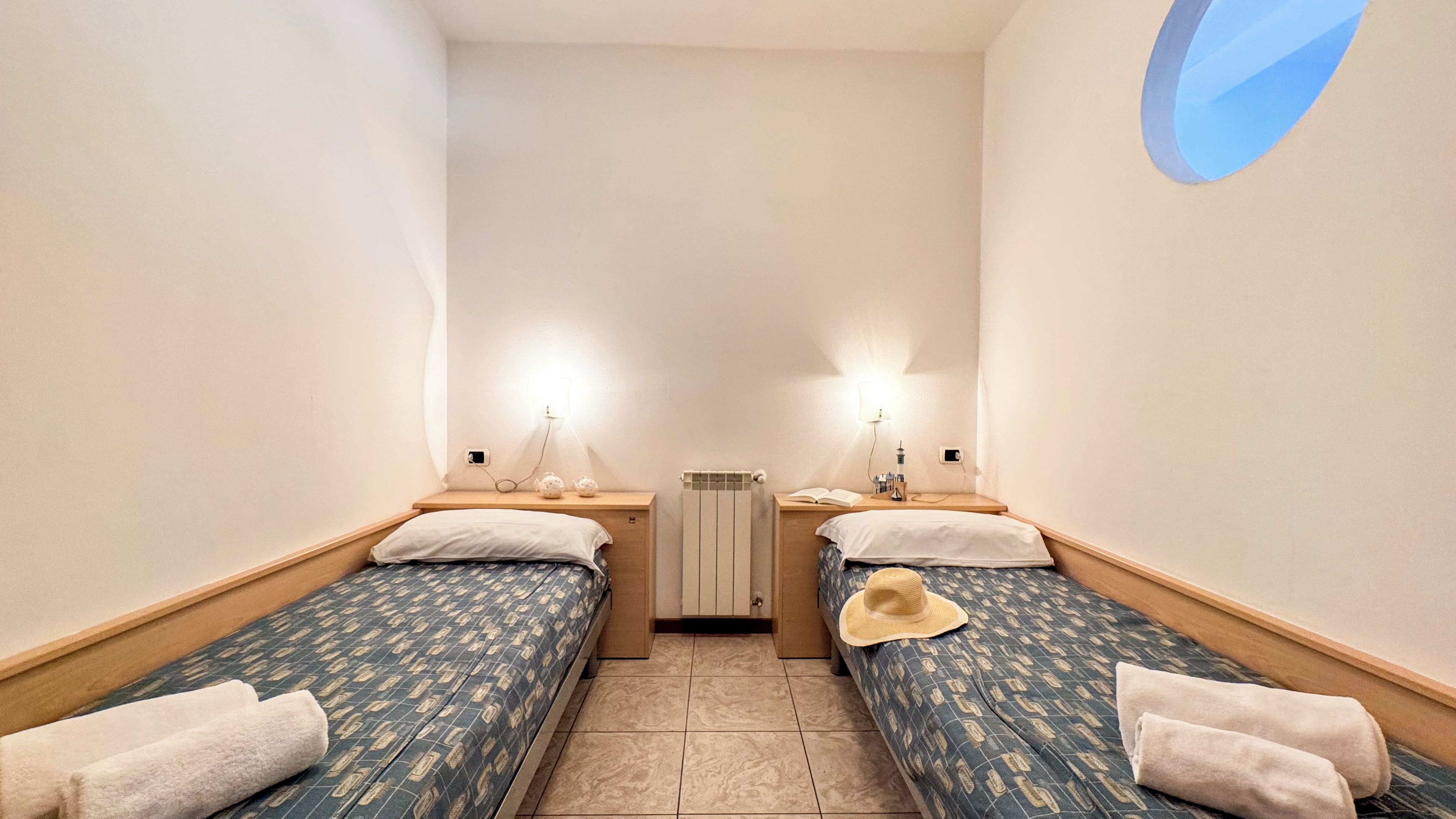 Une petite chambre accueillante avec deux lits simples, chacun muni d’un oreiller et d’une serviette enroulée. Un radiateur se trouve entre les lits, ainsi qu’une petite table sur laquelle sont posés une lampe et un livre.