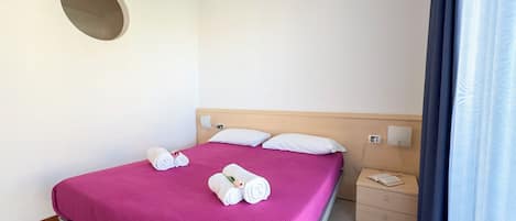 En pænt redt seng med et rosa sengetæppe, to hvide puder og to sammenrullede håndklæder med en blomst. Der er et trænatbord med en bog og en stikkontakt.
