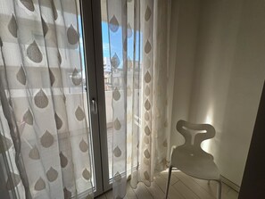 A room with a sliding glass door with a leaf-patterned sheer curtain, a white chair, and a view of buildings outside. - Casa Helios by Ca-sa Check (Chioggia)