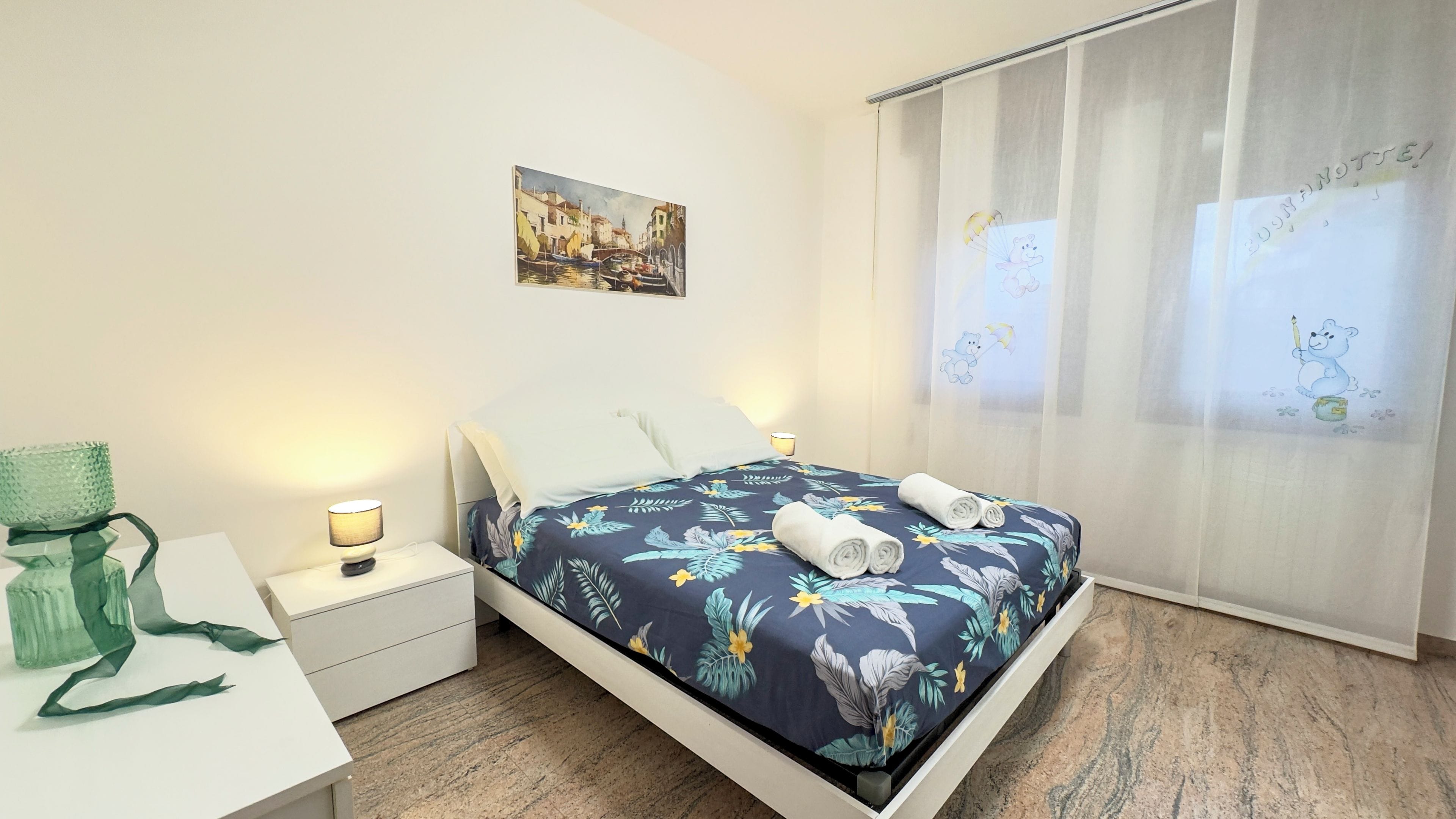 A hotel room with a bed, a nightstand, a lamp, a painting on the wall, and a window with curtains.