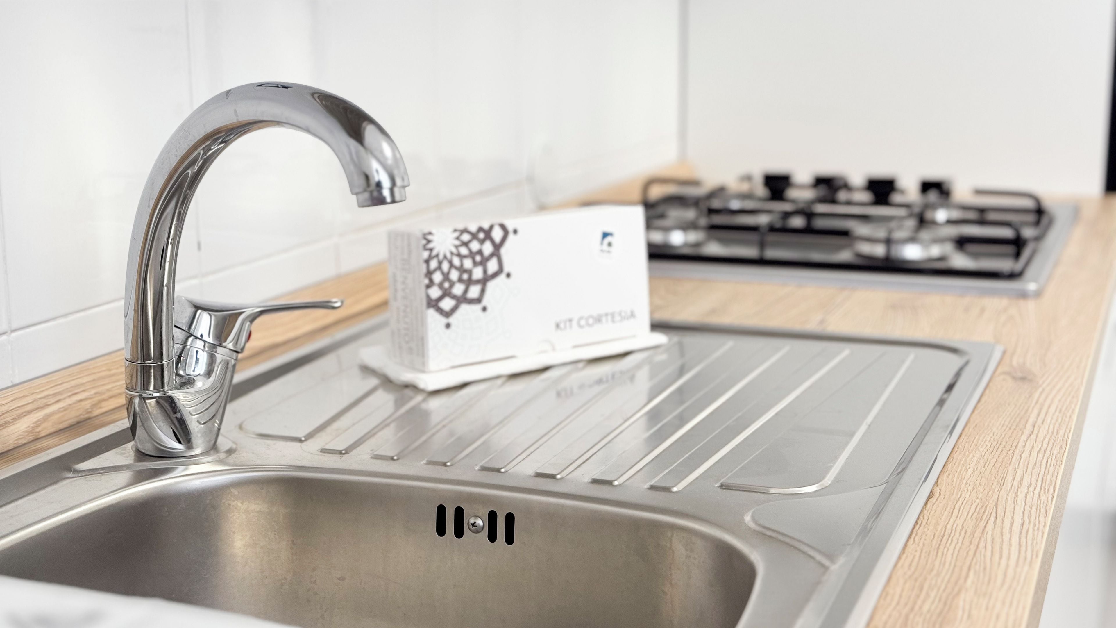 Eine Spüle aus Edelstahl mit modernem Wasserhahn, eine mit 'KIT CONTESIA' beschriftete Box und ein Gaskochfeld im Hintergrund.