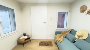 A compact living space with a door, a small bench with a book, a blue sofa with a hat, and a window with a view. - Ca' Egle by Ca-sa Check (Chioggia)