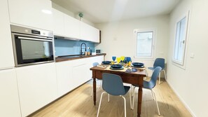 A compact kitchen with white cabinets, a built-in oven, and a microwave. There is a dining table with blue chairs and a small window above it. - Ca' Egle by Ca-sa Check (Chioggia)