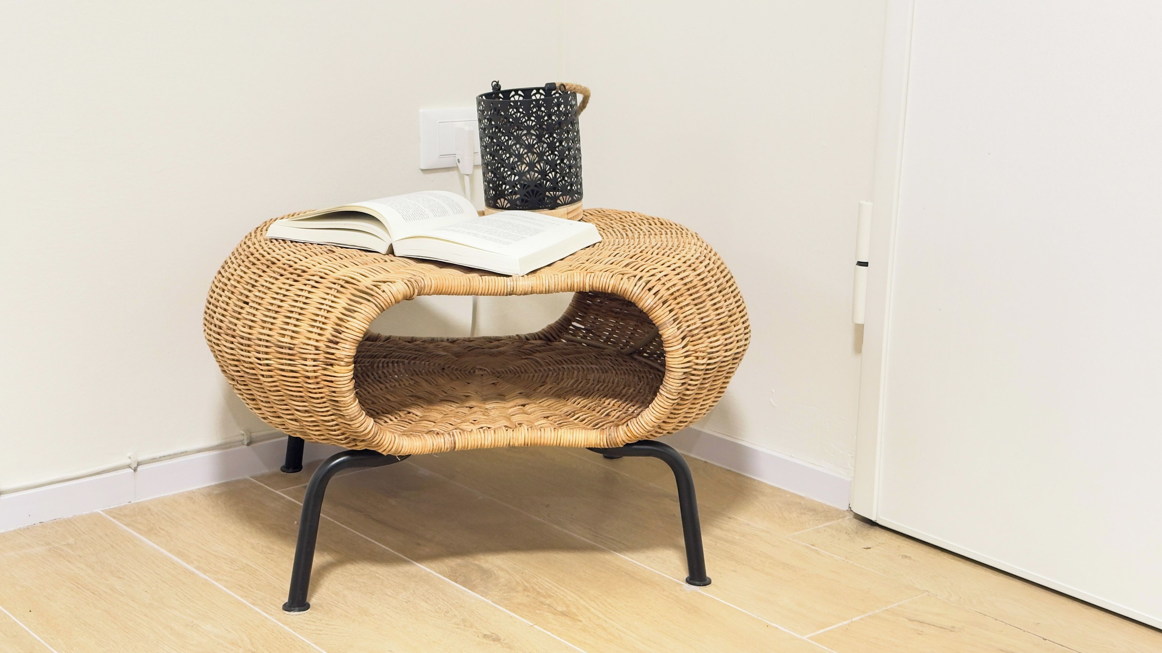 Una silla de mimbre con un libro encima, ubicada en una esquina junto a una pared blanca y suelo de madera.