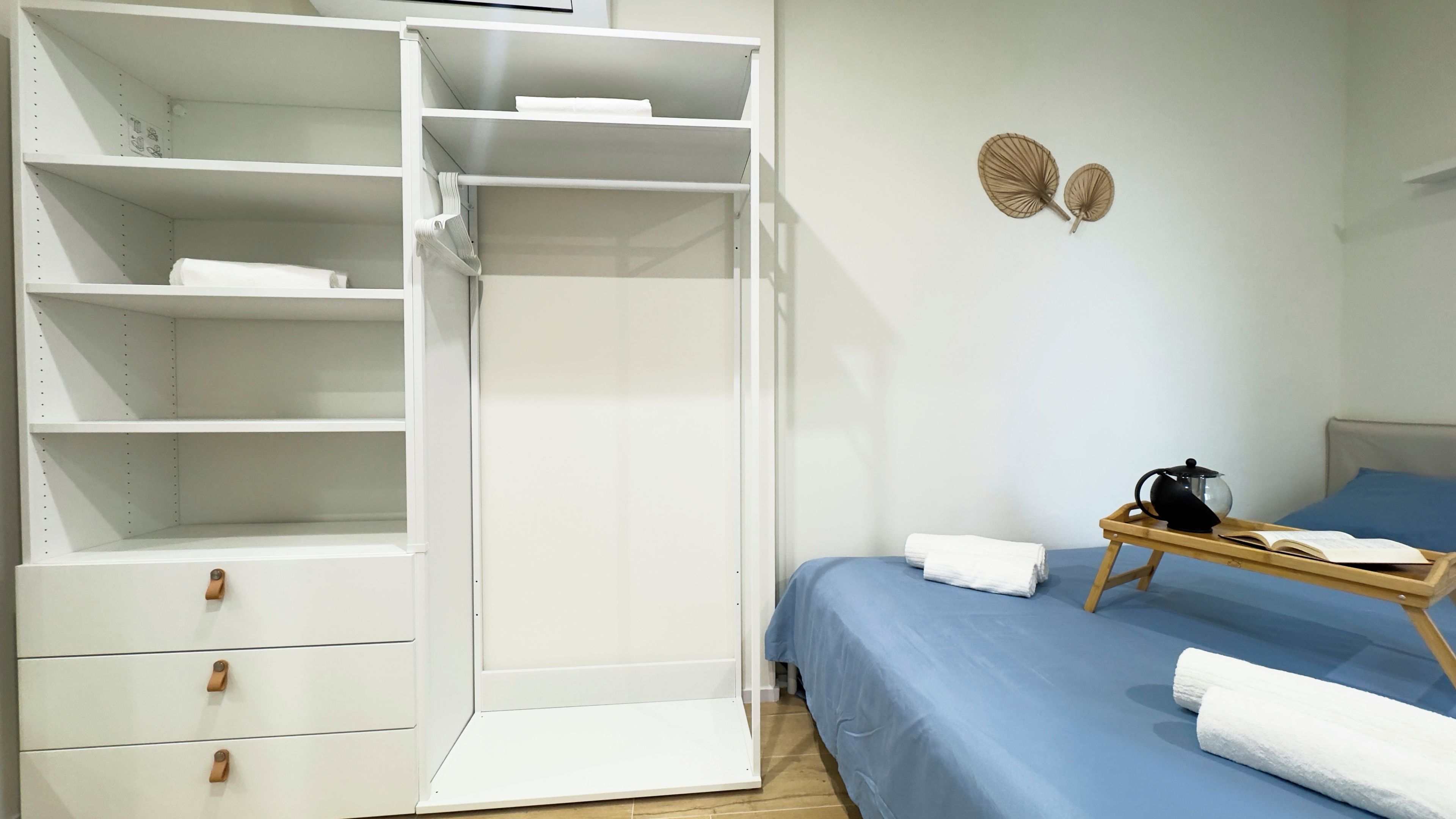 Un dormitorio con un armario blanco, una cama con ropa de cama azul, una mesita de noche de madera con un hervidor y una bandeja con un libro y una taza.