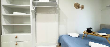 Un dormitorio con un armario blanco, una cama con ropa de cama azul, una mesita de noche de madera con un hervidor y una bandeja con un libro y una taza.