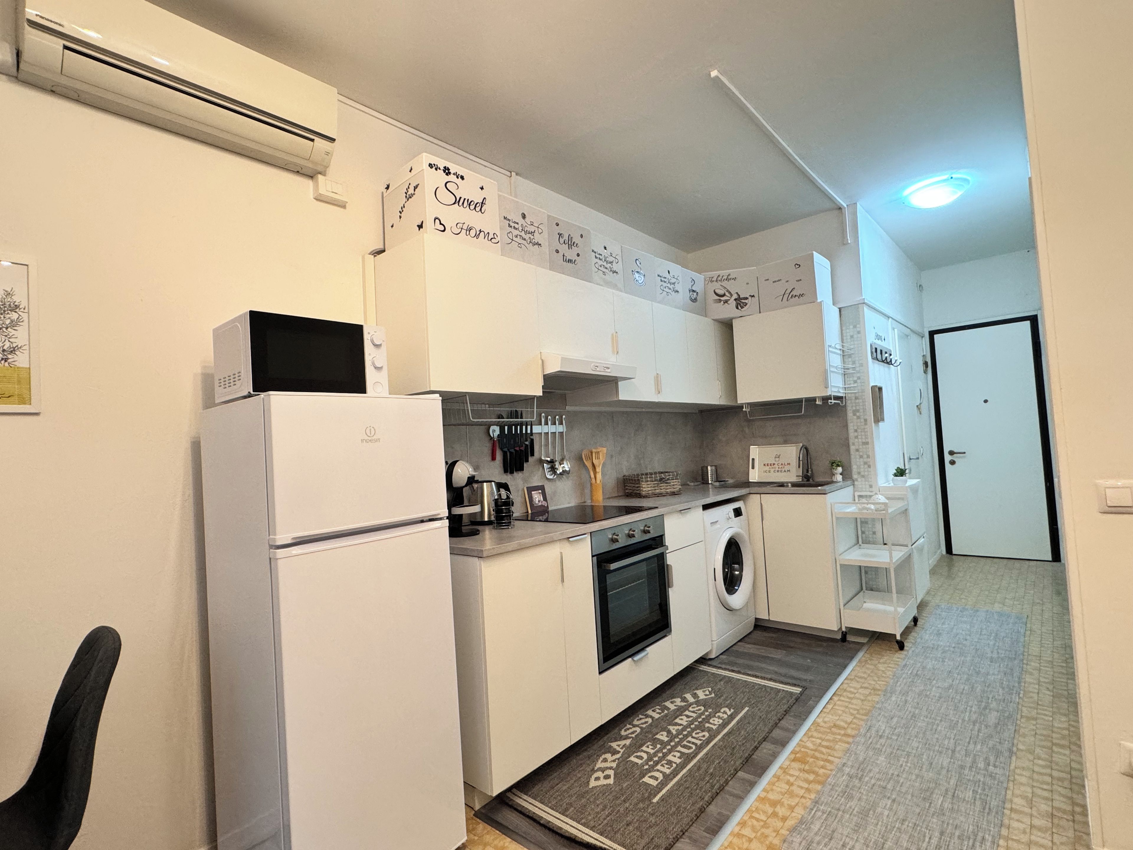 Une cuisine moderne avec des armoires blanches, un four encastré, un four à micro-ondes et une machine à café. On y trouve un petit évier et un réfrigérateur. La cuisine est décorée dans des tons de gris et de blanc, avec des panneaux décoratifs sur le mur.