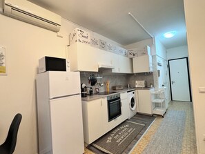 Une cuisine moderne avec des armoires blanches, un four encastré, un four à micro-ondes et une machine à café. On y trouve un petit évier et un réfrigérateur. La cuisine est décorée dans des tons de gris et de blanc, avec des panneaux décoratifs sur le mur.