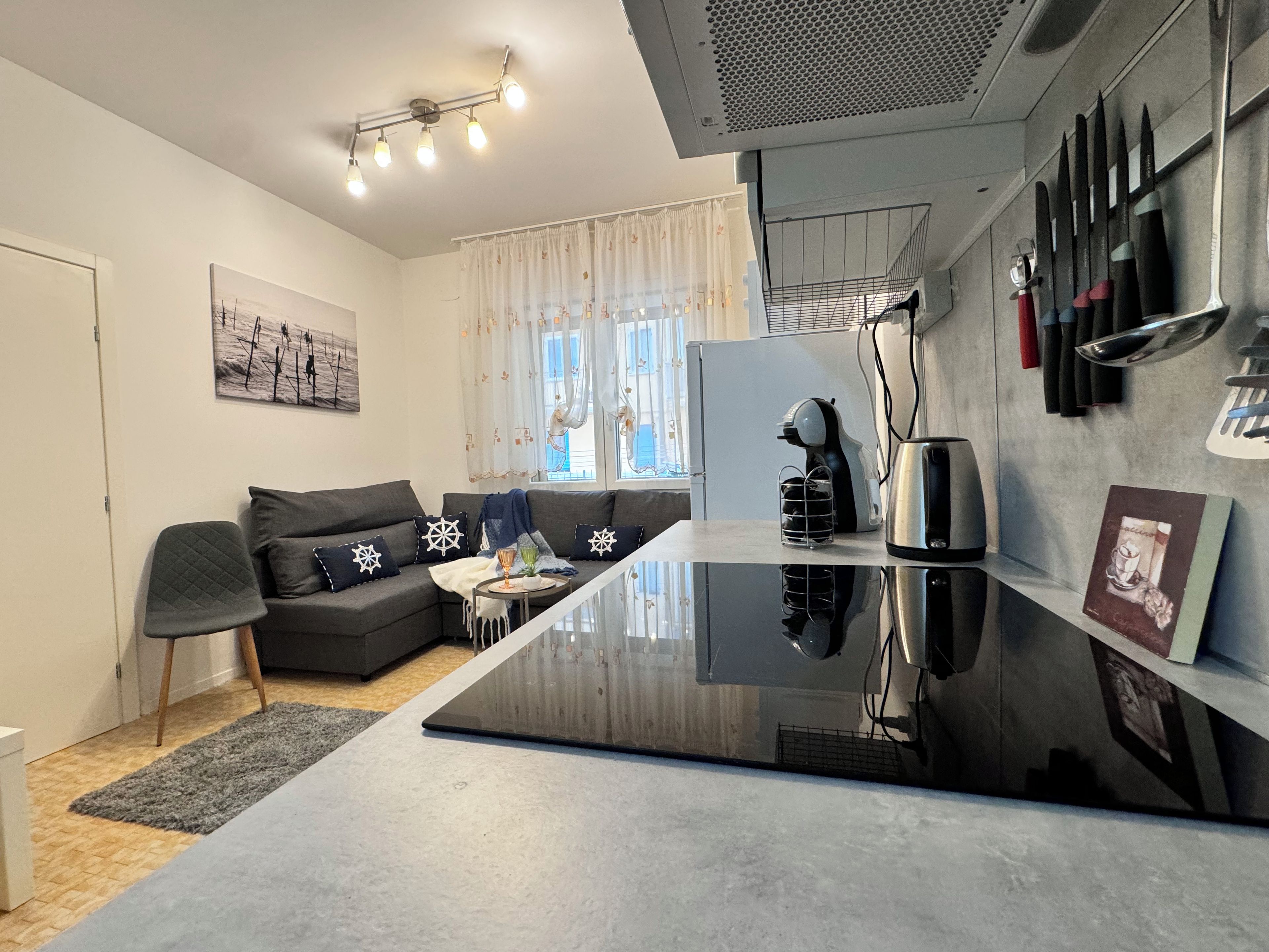 A modern kitchen with a black countertop, a built-in oven, and a coffee machine. There is a grey sofa with star-patterned cushions and a small table with a glass and a cup.