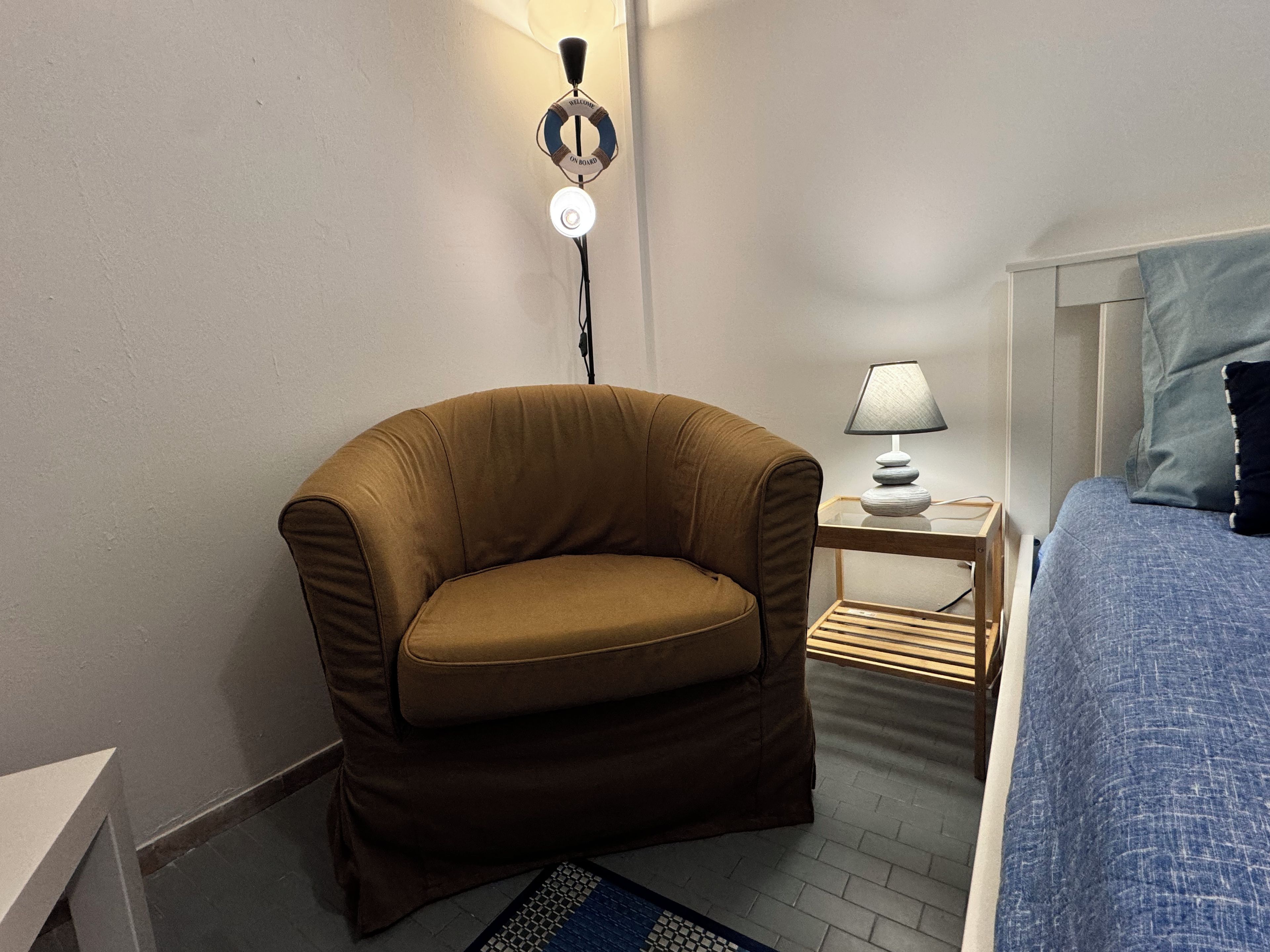 A small, cozy room with a brown armchair, a bedside table with a lamp, and a bed with a blue blanket.