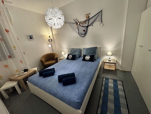 Un dormitori amb llit, cadira, tauleta de nit, làmpada i un objecte decoratiu penjant.