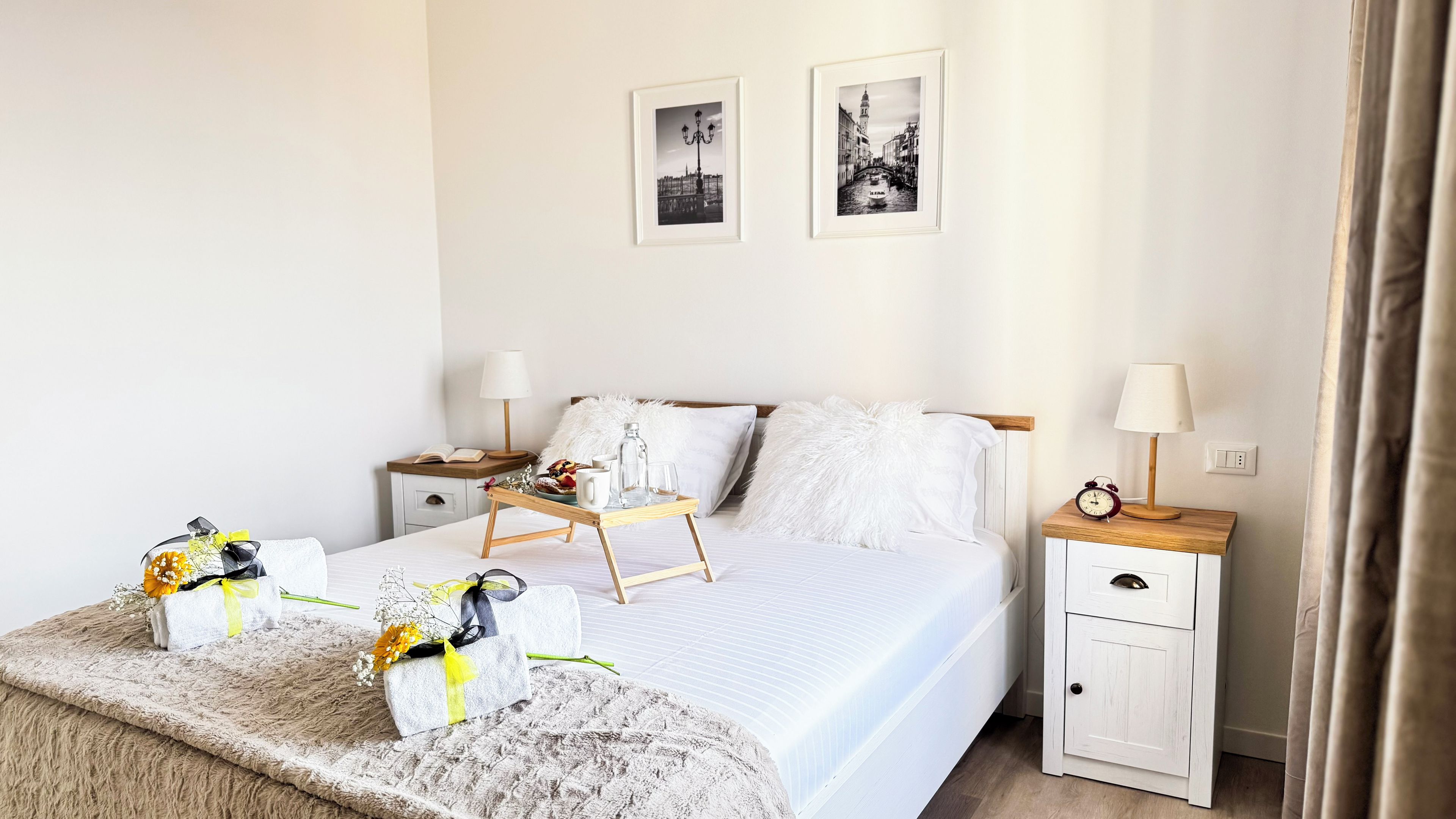 A neatly arranged hotel room with a large bed, two bedside tables, a bedside lamp, a small clock, and a framed picture on the wall.