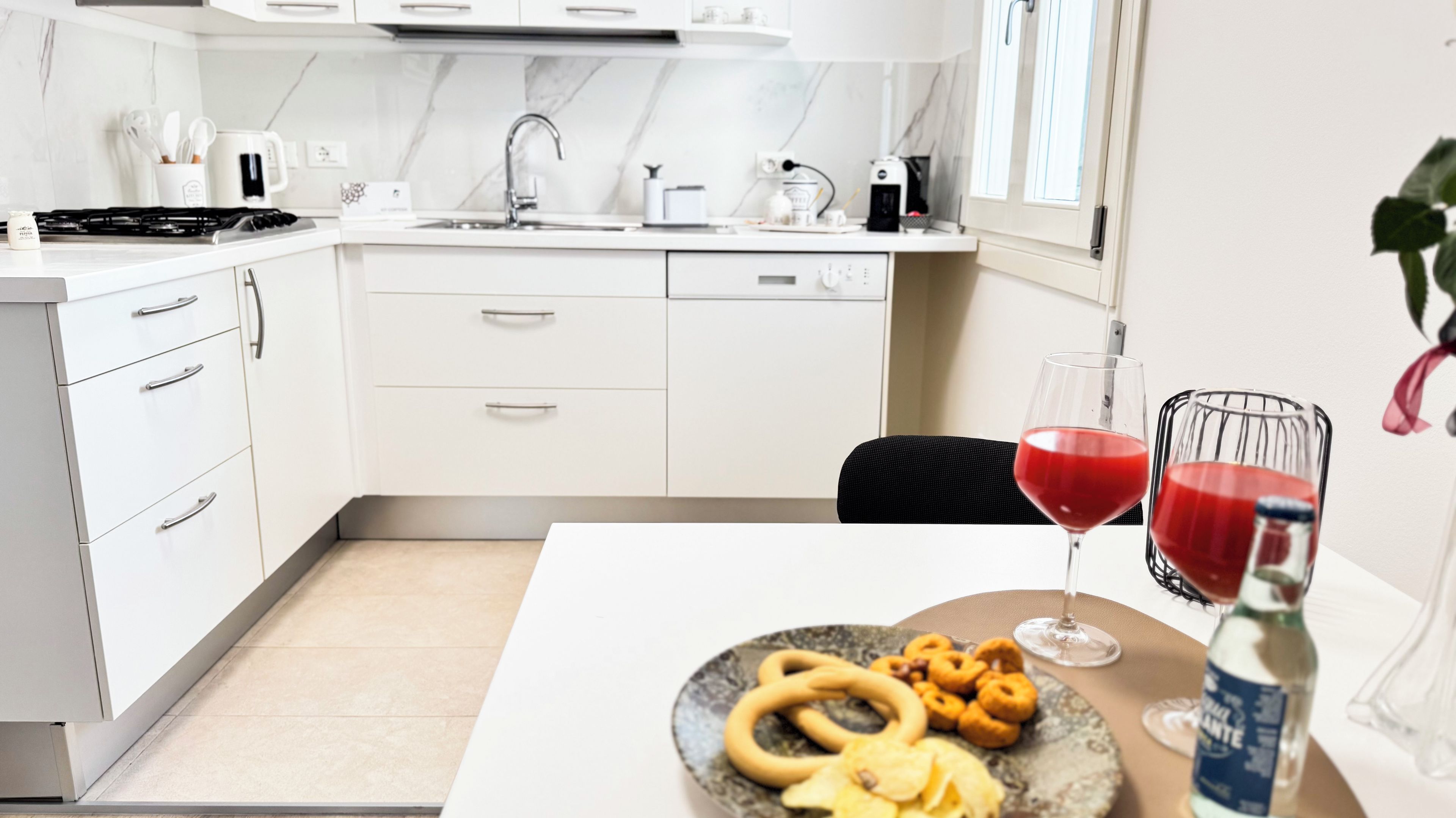 A modern kitchen with white cabinets, a stove, and a sink. There is a dining table with a plate of snacks and two glasses of red drink.