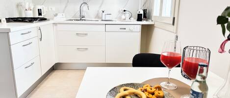 A modern kitchen with white cabinets, a stove, and a sink. There is a dining table with a plate of snacks and two glasses of red drink.