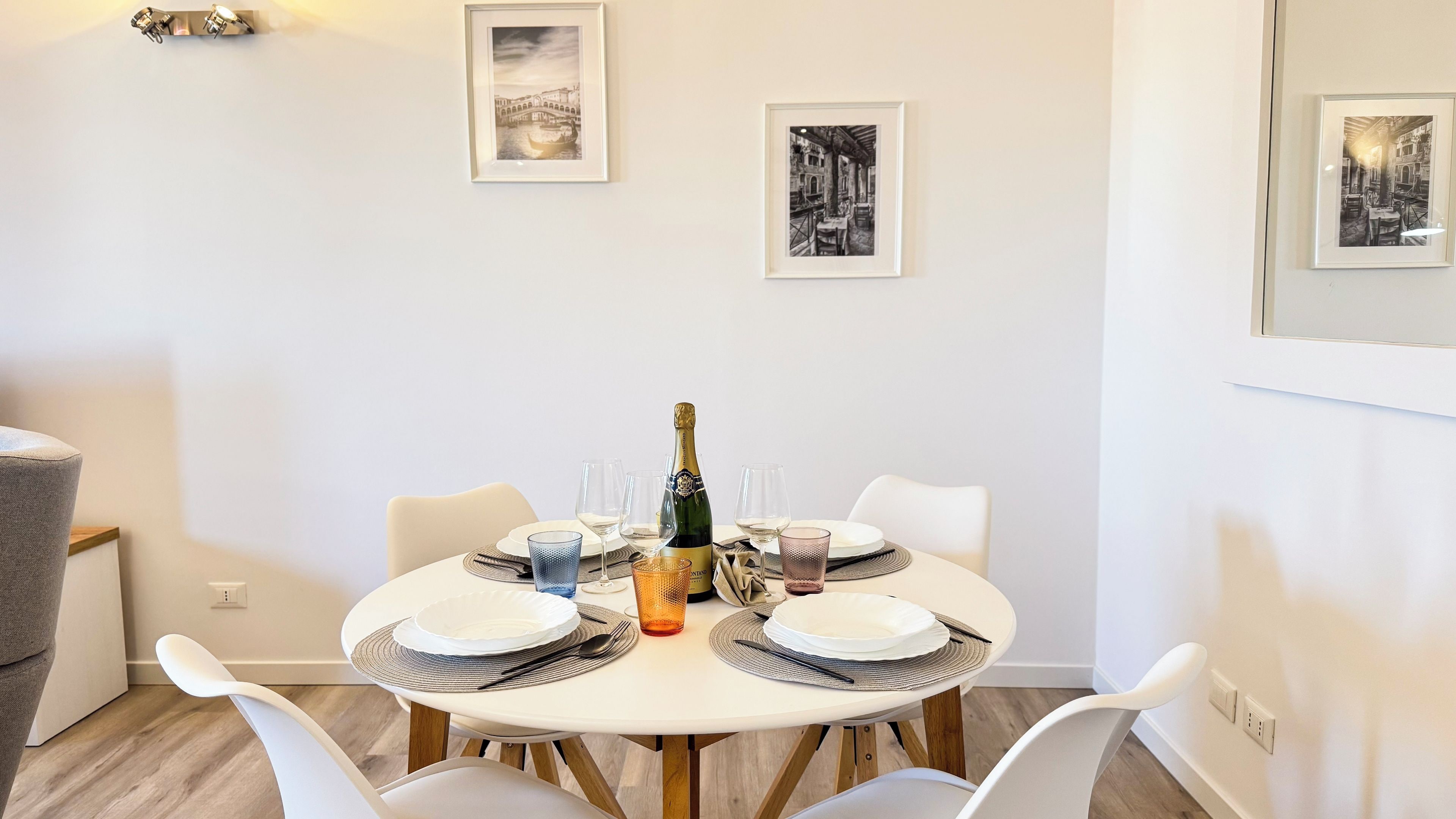 A dining area with a round table set for four, featuring a bottle of champagne, white plates, and glasses. The room has a minimalist design with white walls and a framed picture above the table.