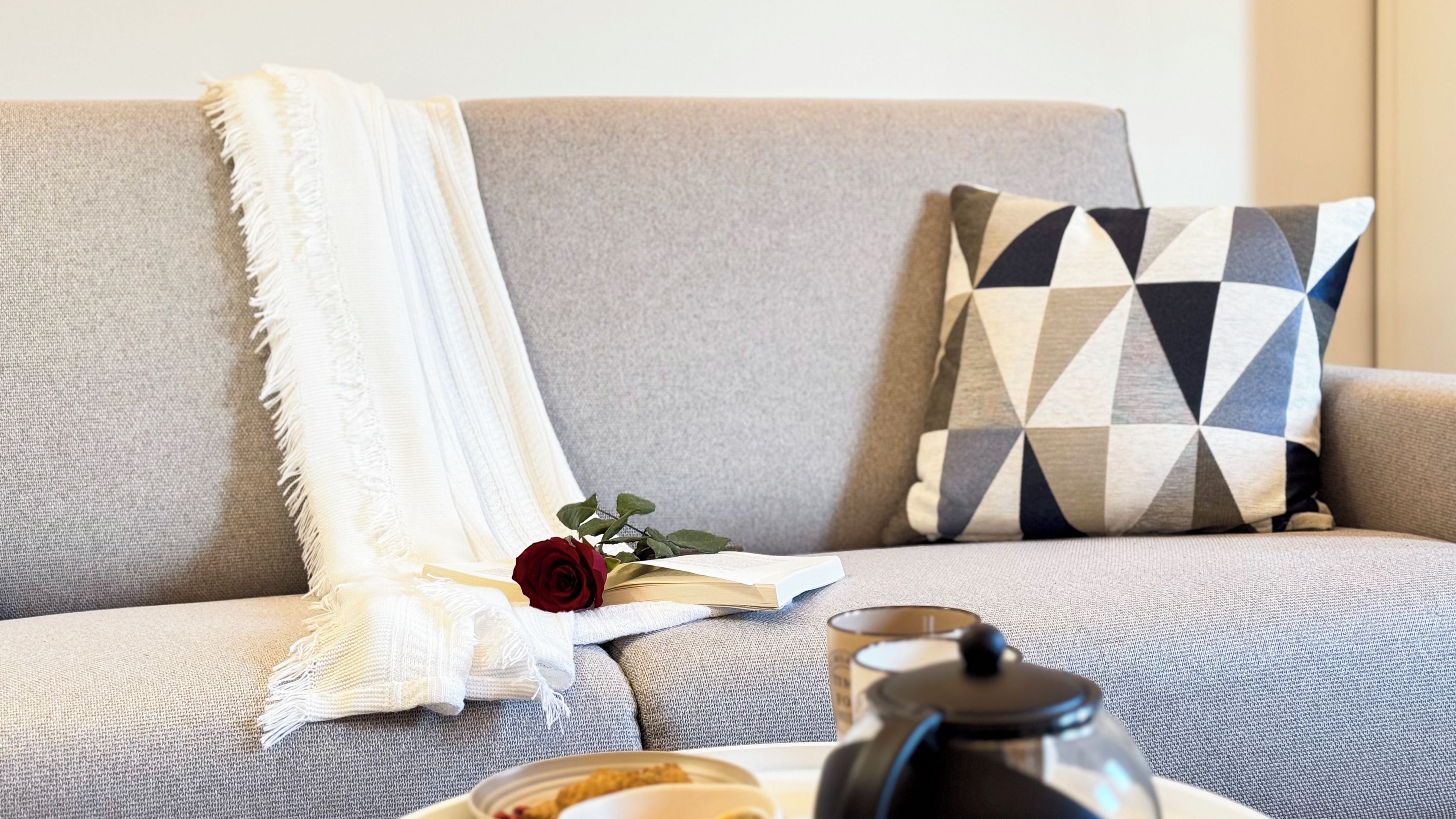A grey sofa with a white throw blanket and a geometric patterned pillow. A red rose is placed on the sofa, and a teapot is on a table in front of it.