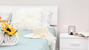 Uma cama bem arrumada com um travesseiro branco e fofo, uma toalha branca enrolada e um chapéu branco sobre um cobreleito azul claro.
