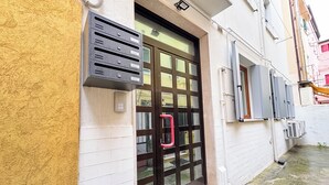 Entrada de edifício com abertura para correspondência em metal preto, porta de vidro com puxador vermelho e um pequeno degrau que leva à porta.