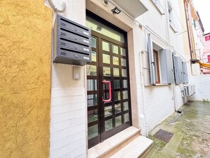 Entrada de edificio con buzón de metal negro, puerta de vidrio con manija roja y un pequeño escalón que lleva a la puerta.