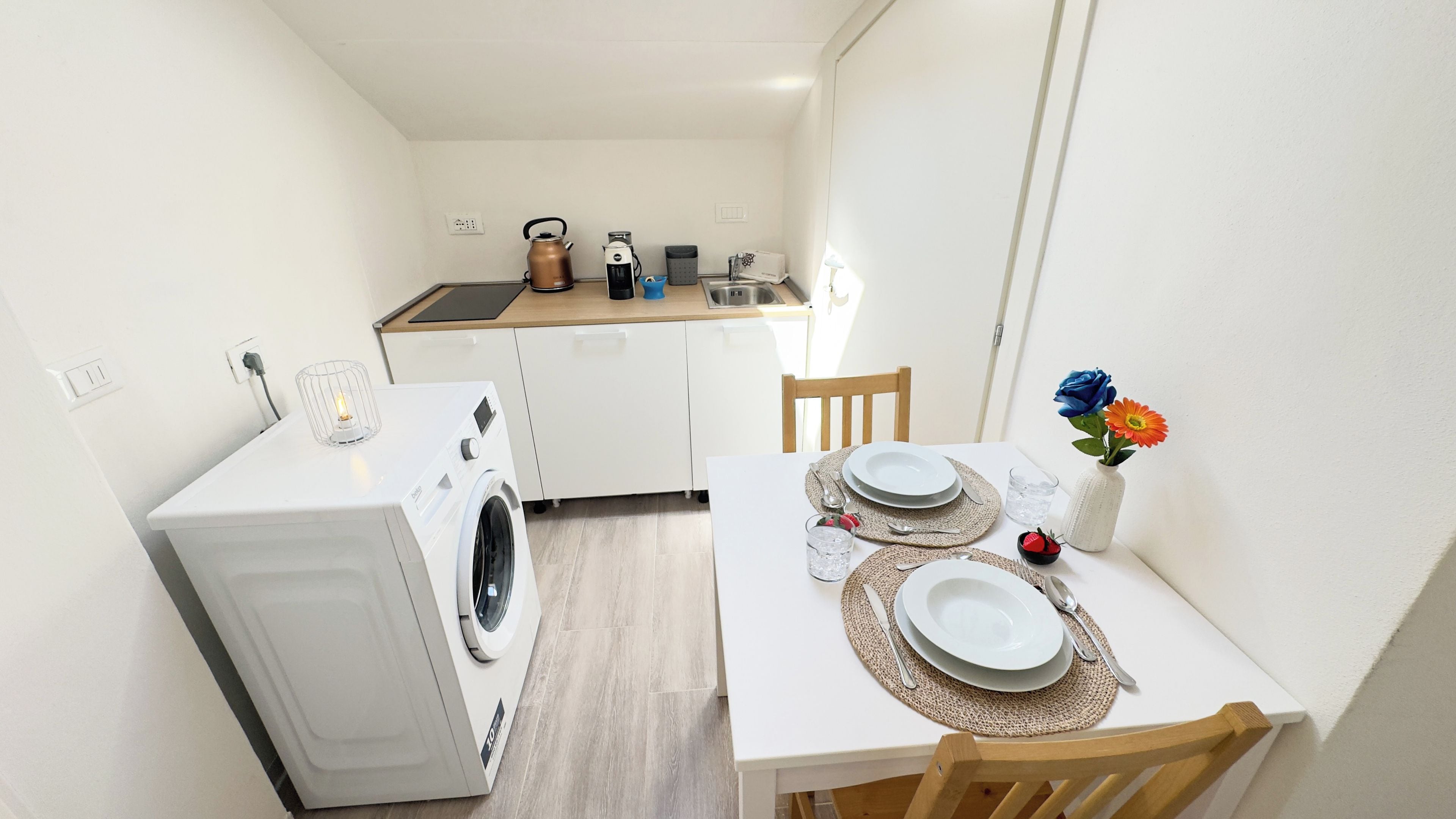 Une cuisine compacte équipée d’un lave-linge, d’une bouilloire et d’un grille-pain. Une table à manger est dressée, avec des assiettes et un vase de fleurs.