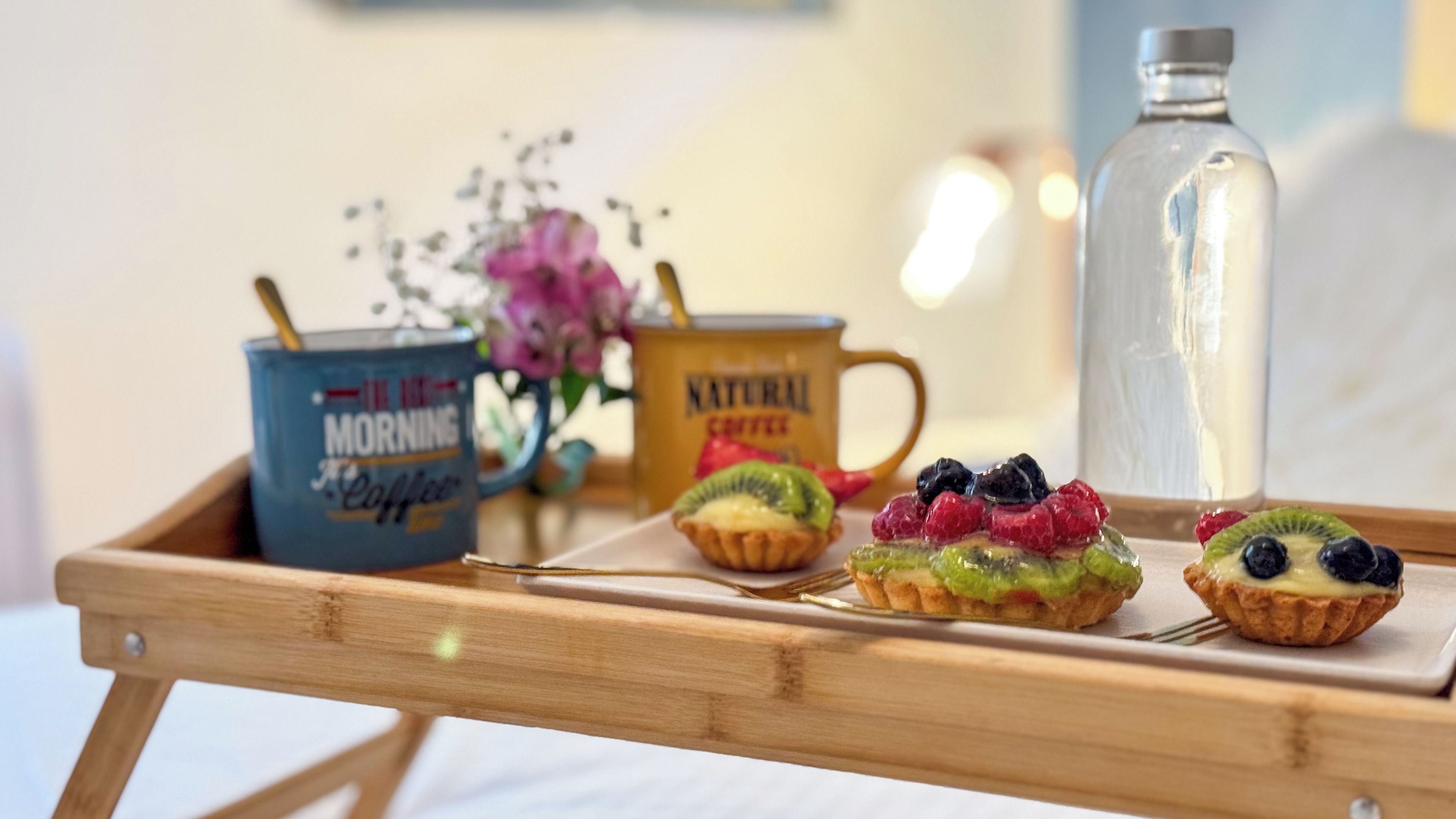 A wooden tray with three fruit tarts and two coffee mugs on a table.