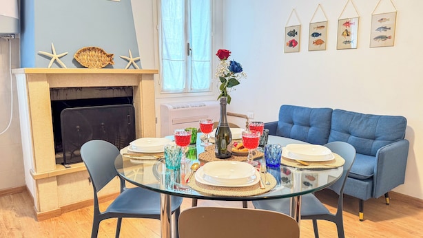 A dining area with a glass table set for four, surrounded by blue chairs. A blue sofa is against the wall, and a fireplace with starfish decorations is visible. - L'ultimo Venchero by Ca-sa Check (Chioggia)
