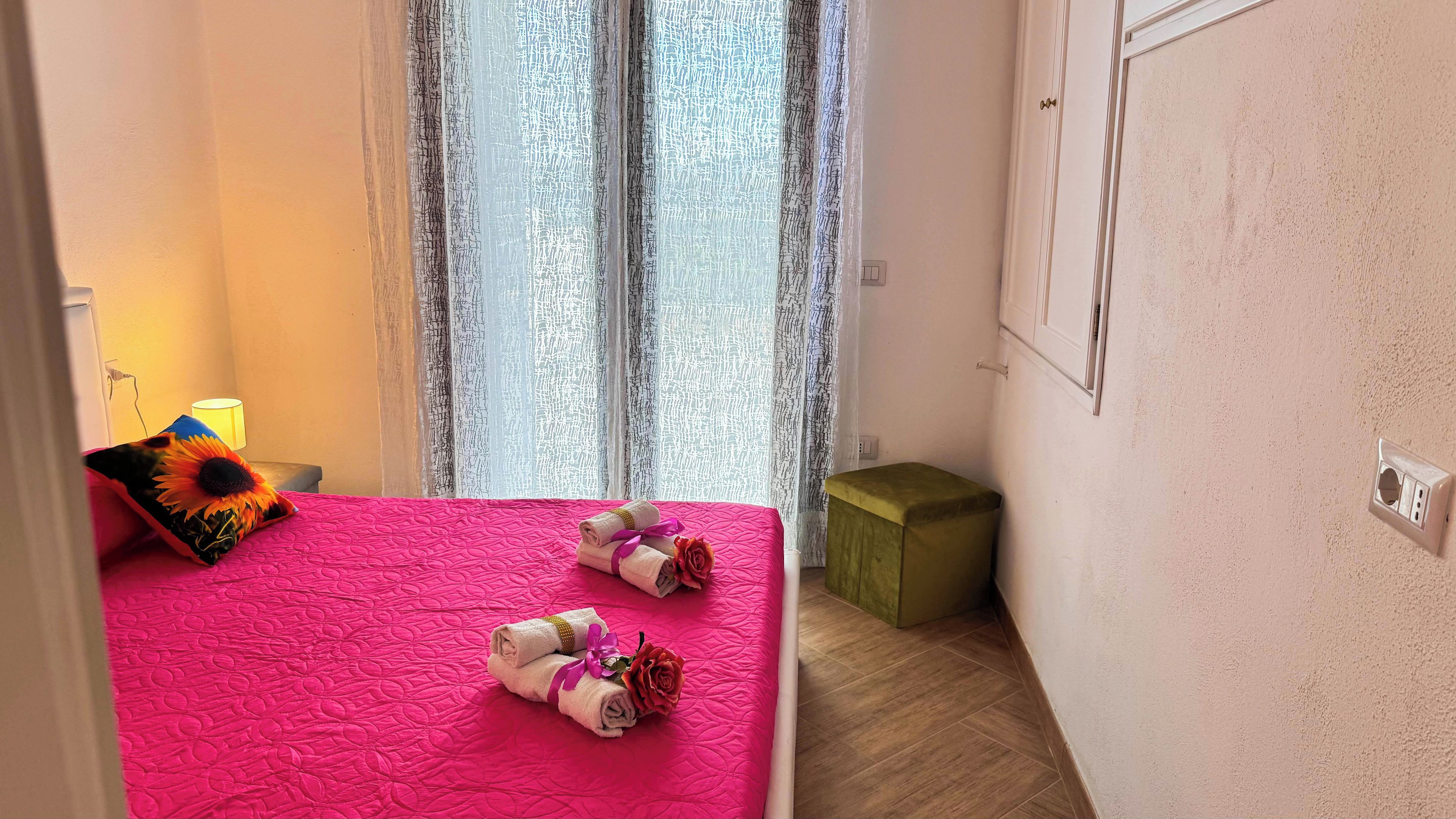 A room with a pink bedspread, a small bedside table with a lamp, and a green trash bin.