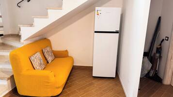 A compact living space with a white refrigerator, a yellow sofa with patterned cushions, and a staircase leading to an upper level.