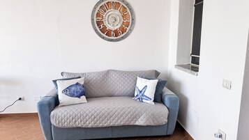 A room with a grey quilted sofa, two cushions with fish and starfish designs, and a wooden clock on the wall.