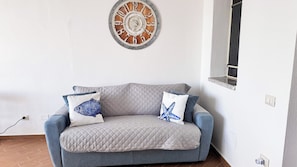 A room with a grey quilted sofa, two cushions with fish and starfish designs, and a wooden clock on the wall. - La Casa Delle Stelle by Ca-sa Check (San Teodoro)