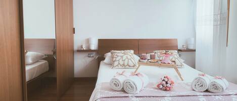 Habitación de hotel con una cama de madera, una mesita de noche con lámpara, una ventana con cortinas traslúcidas y un mueble auxiliar.