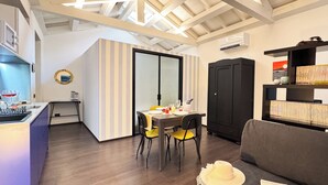 A modern kitchen with a built-in stove, a dining area with a table and chairs, a black wardrobe, and a bookshelf with books. - Tema Apartment by Ca-sa Check (Chioggia)