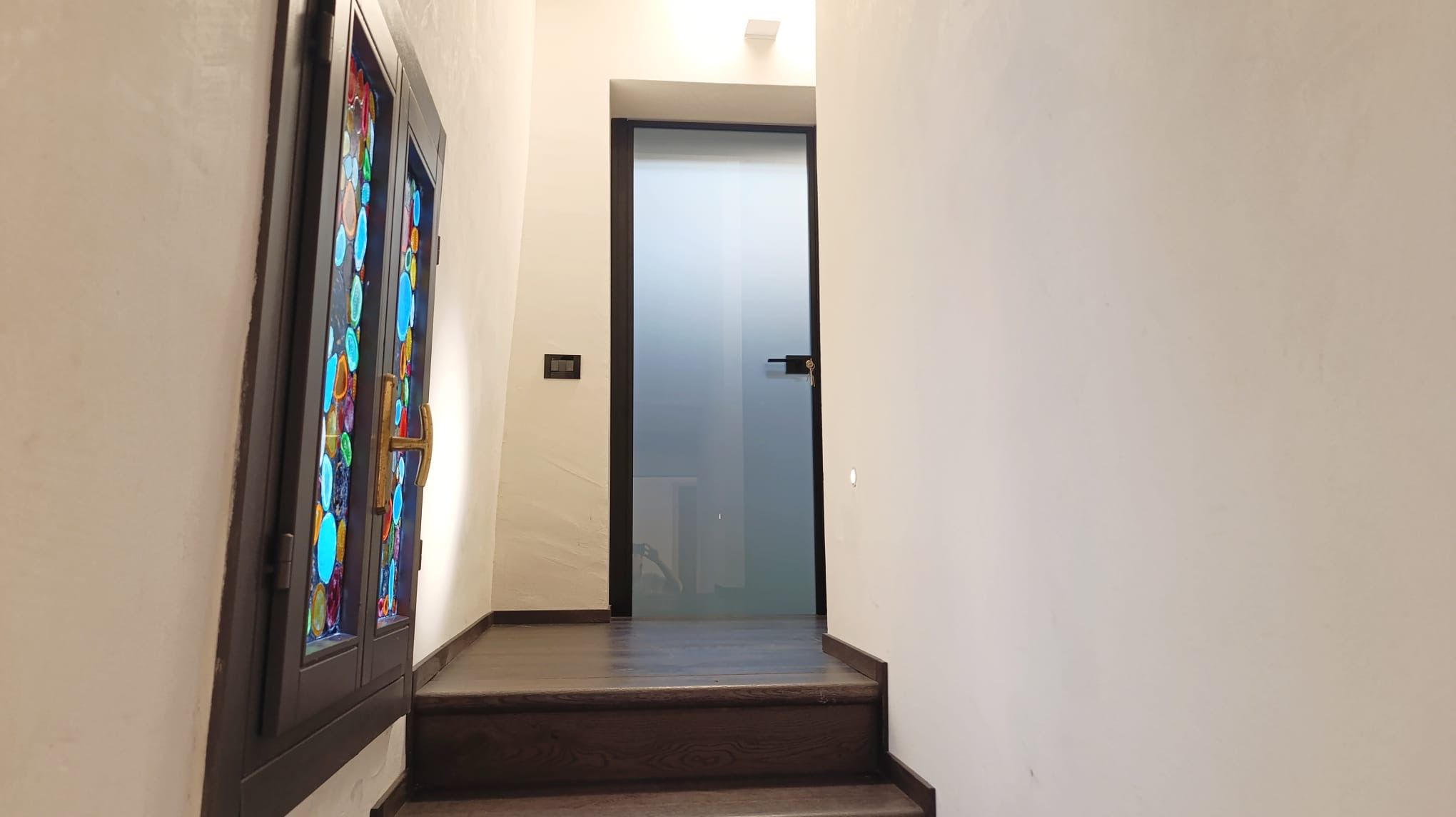 Un pasillo estrecho con una escalera de madera que conduce a una puerta de cristal satinado.