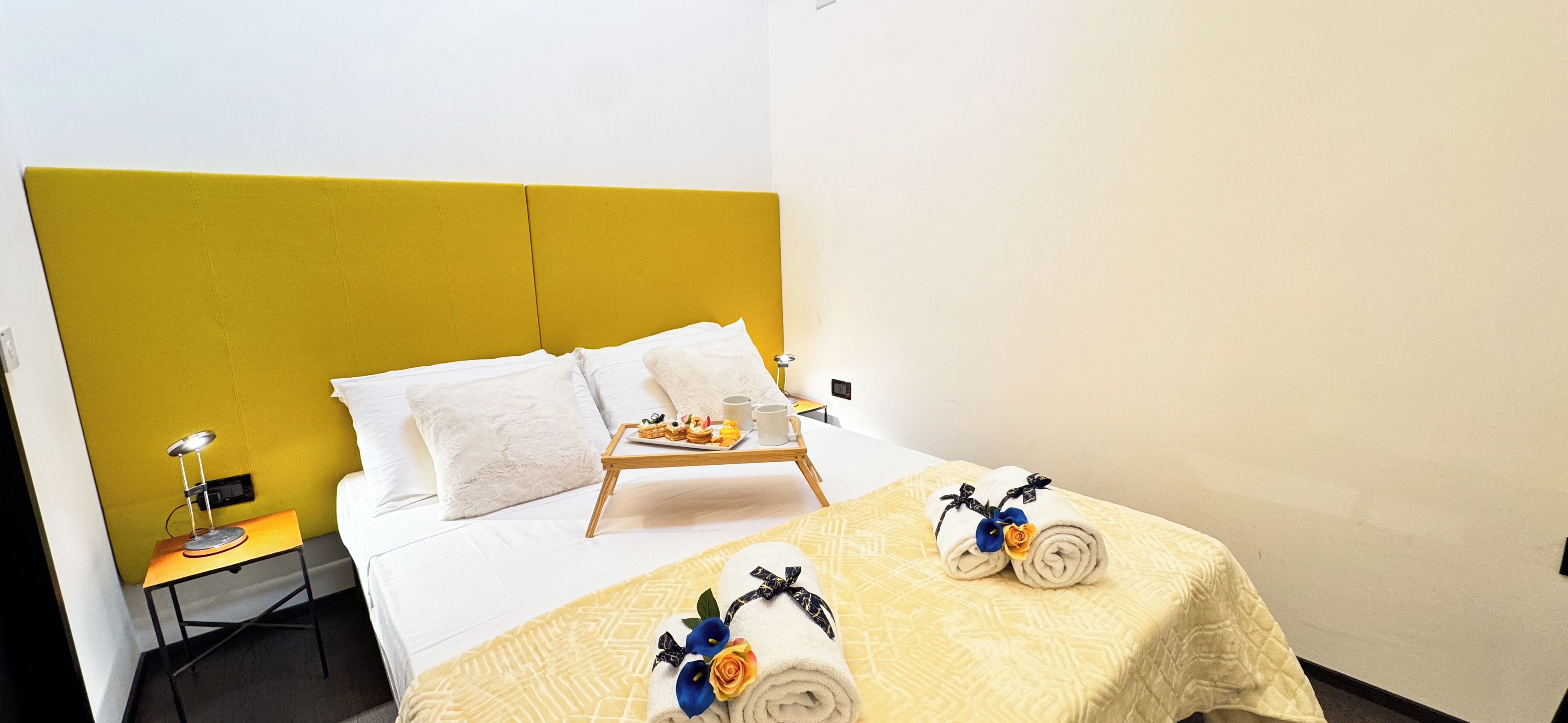 Habitación de hotel con cabecera amarilla, ropa de cama blanca, mesita de noche con lámpara y toallas con adornos florales.