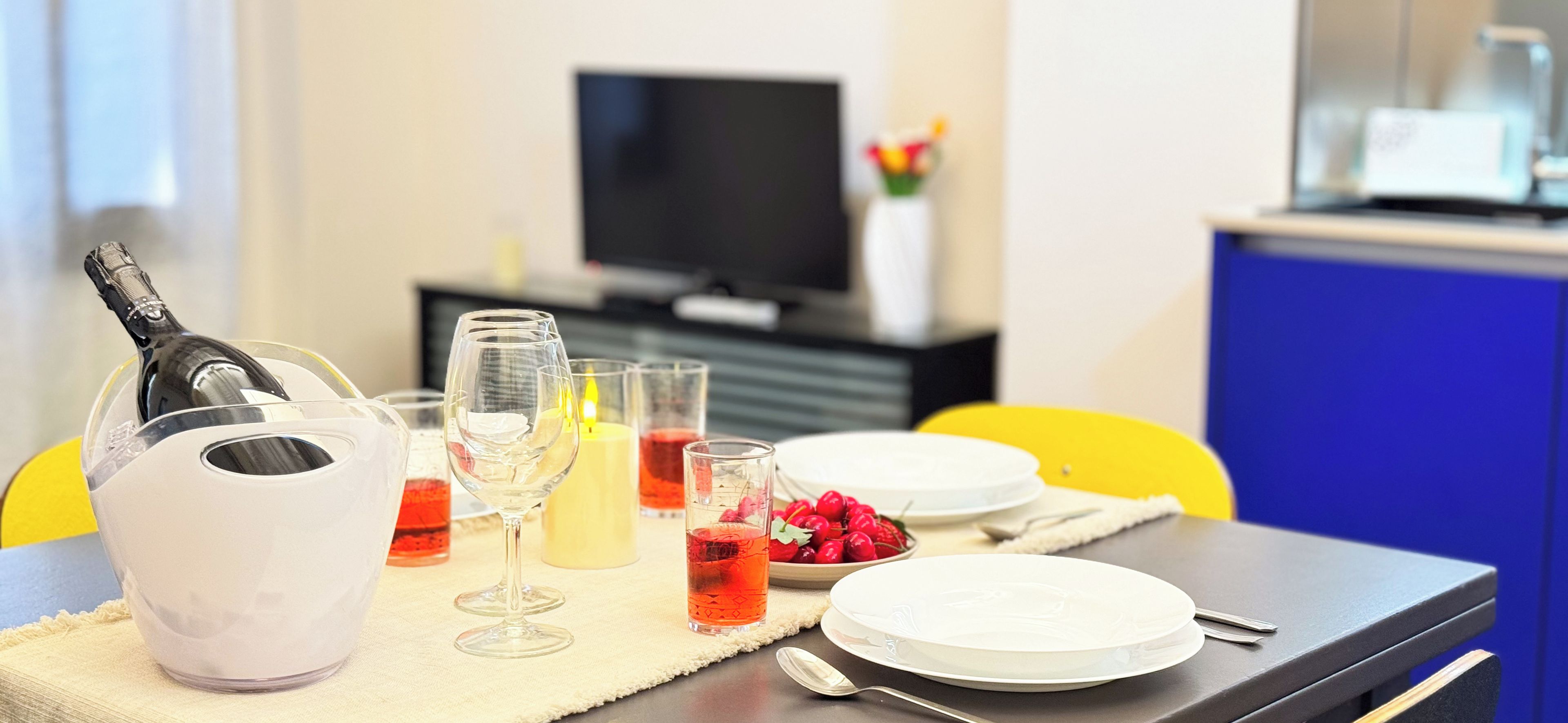 A dining table set with a bottle, glasses, and plates, with a TV and a cabinet in the background.
