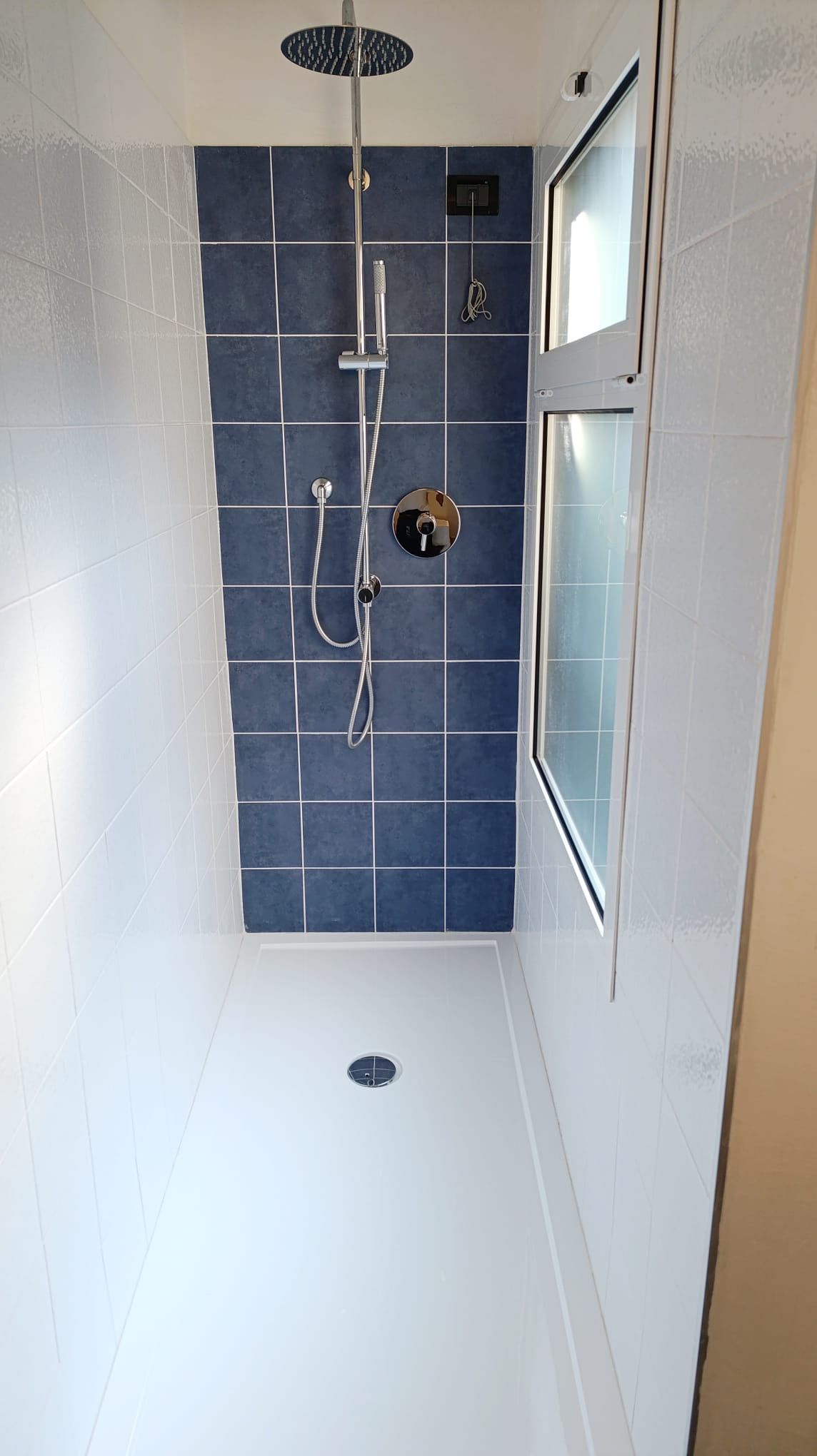 A shower with a handheld showerhead and a wall-mounted faucet, a small control panel, and a drain in the floor.