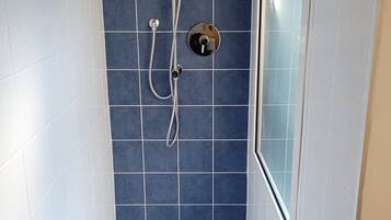A shower with a handheld showerhead and a wall-mounted faucet, a small control panel, and a drain in the floor.