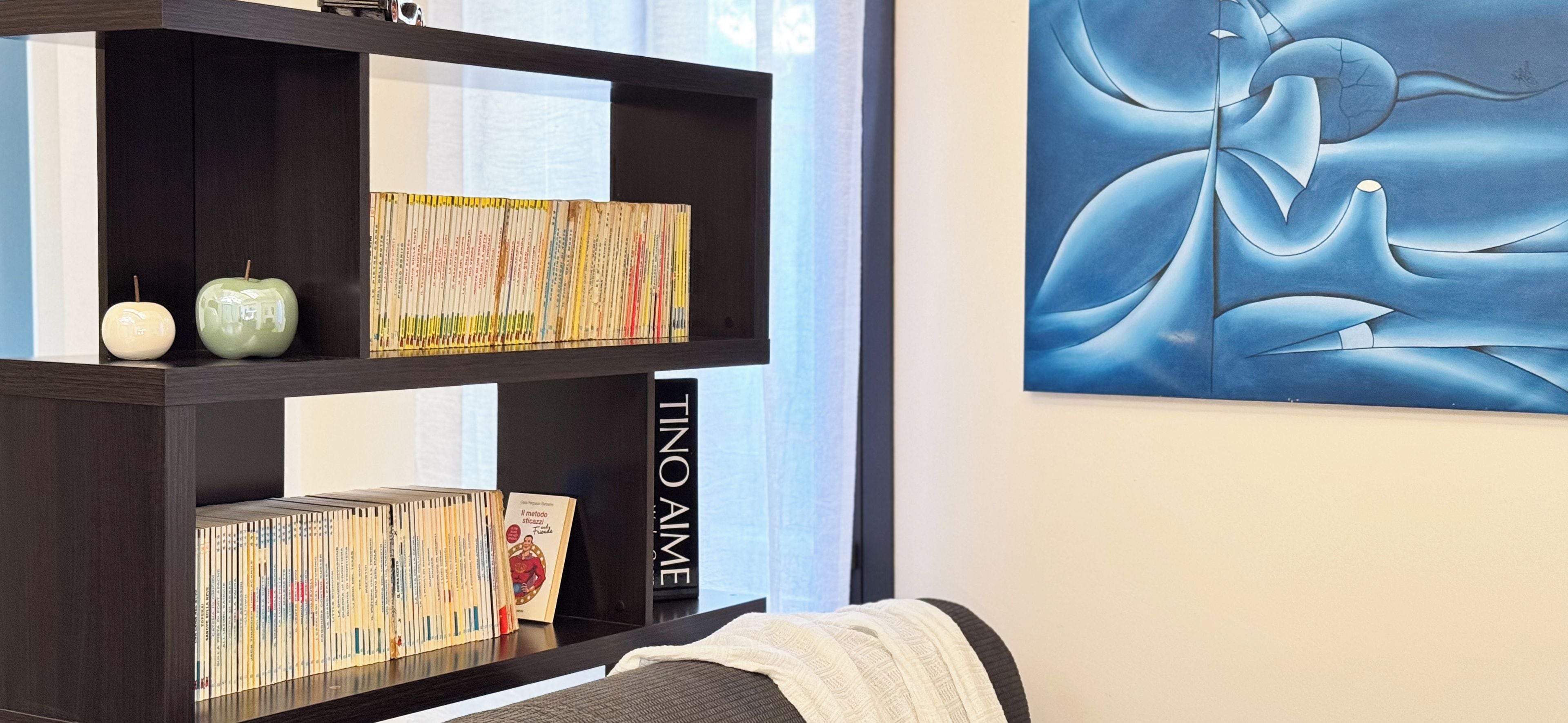 Una estantería con libros y objetos decorativos, un cuadro abstracto enmarcado, y un sofá con una manta.