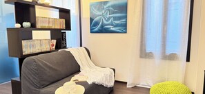 Una sala de estar con sofá, estantería, un cuadro y una ventana con cortinas.