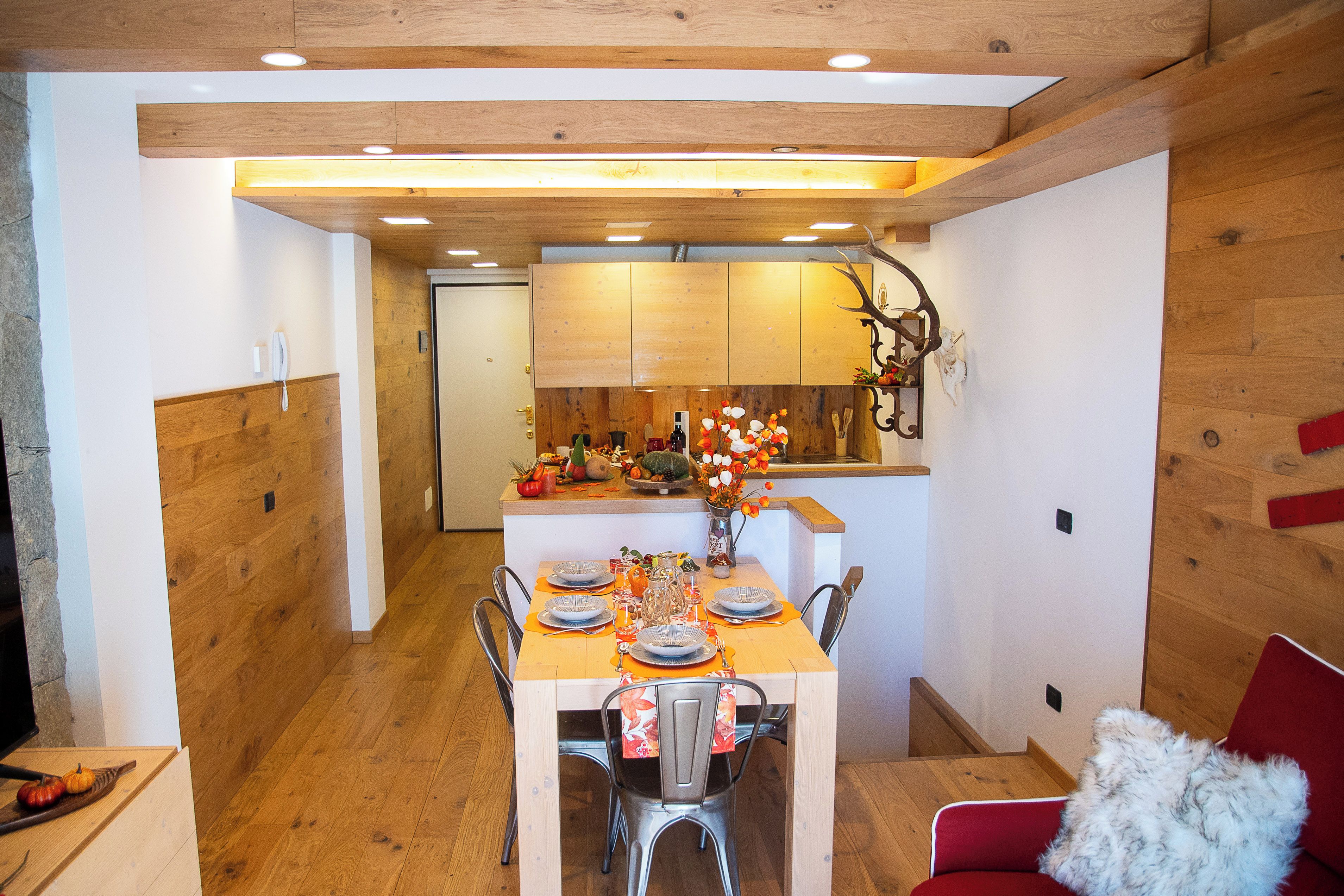 A dining area with a wooden table set for four, surrounded by chairs. A kitchen is visible in the background with wooden cabinets and a countertop.