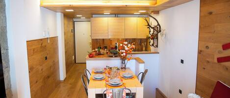 A dining area with a wooden table set for four, surrounded by chairs. A kitchen is visible in the background with wooden cabinets and a countertop.