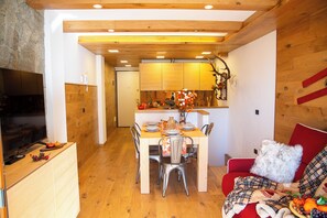 A cozy dining area with a wooden table set for four, surrounded by metal chairs. A red sofa with a plaid blanket is visible in the foreground. - Appartamento Neve by Ca-sa Check (Aprica)