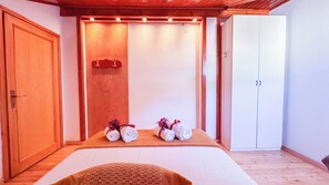 Une pièce avec un plancher en bois, un lit avec un matelas blanc, un banc en bois avec des serviettes et des fleurs, et une armoire blanche.